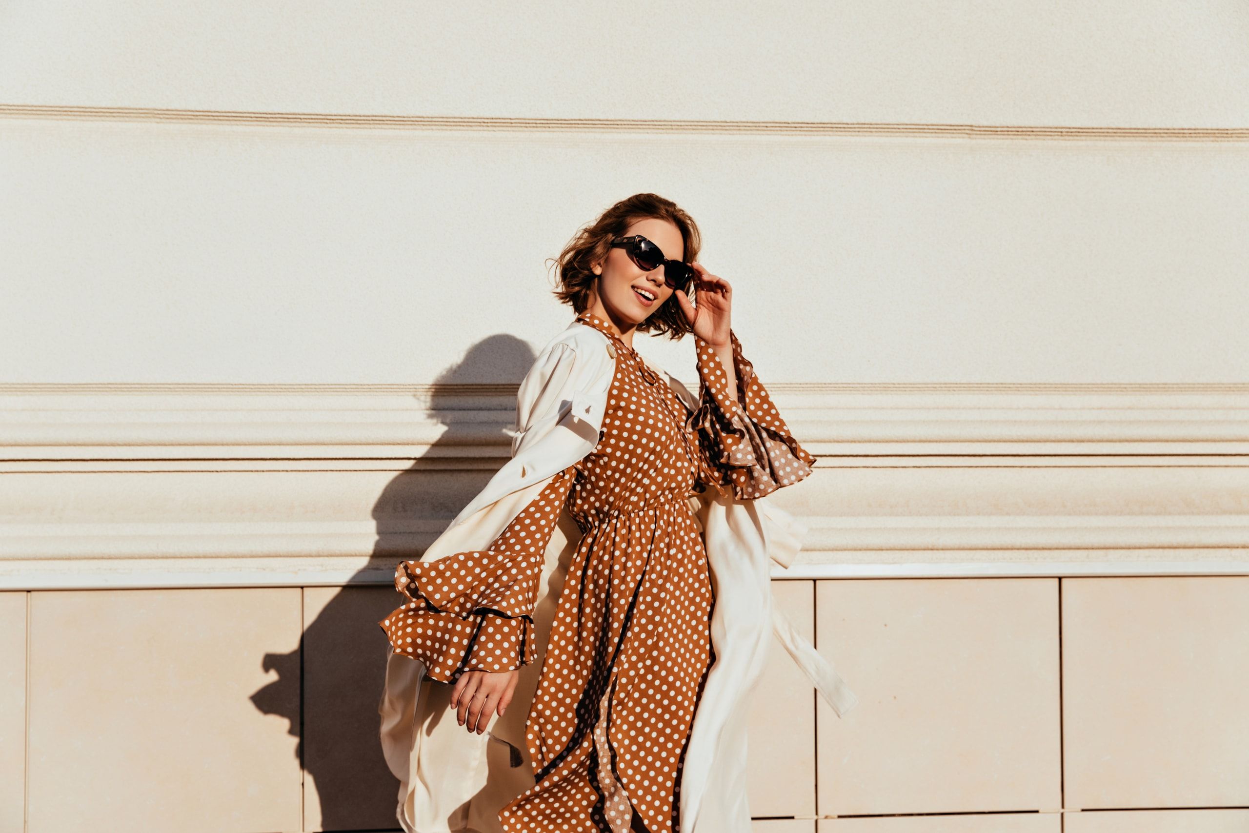 Smiling woman in sunglasses and a rust polka-dot dress with a cream trench coat, walking past a sunlit wall