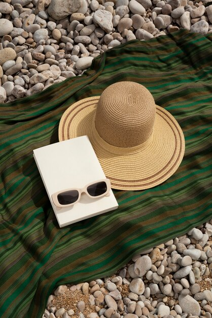 Straw sun hat, book and cream sunglasses on a green striped blanket over pebbles